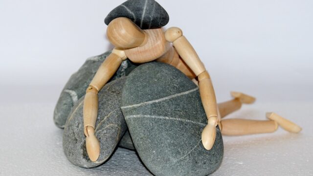 prone wooden figure lying on stones with a stone on its back