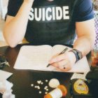 A police officer wearing a buleet proof vest that reads Suicide, sat at a table writing a contract with a gun some pills.