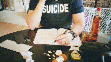 A police officer wearing a buleet proof vest that reads Suicide, sat at a table writing a contract with a gun some pills.